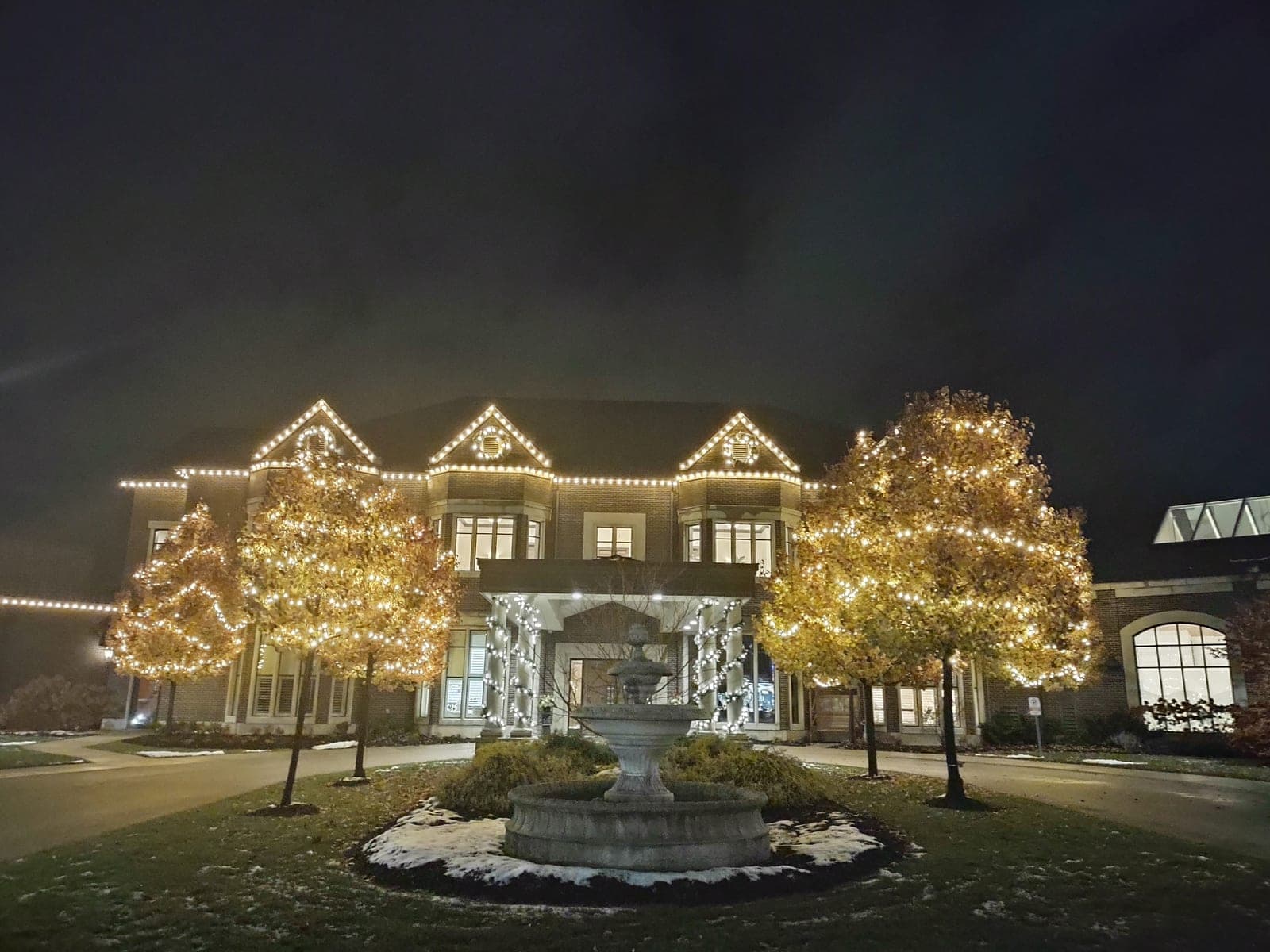 Grand event venue with warm-white roofline Christmas lights and tree wrapping around a stone fountain