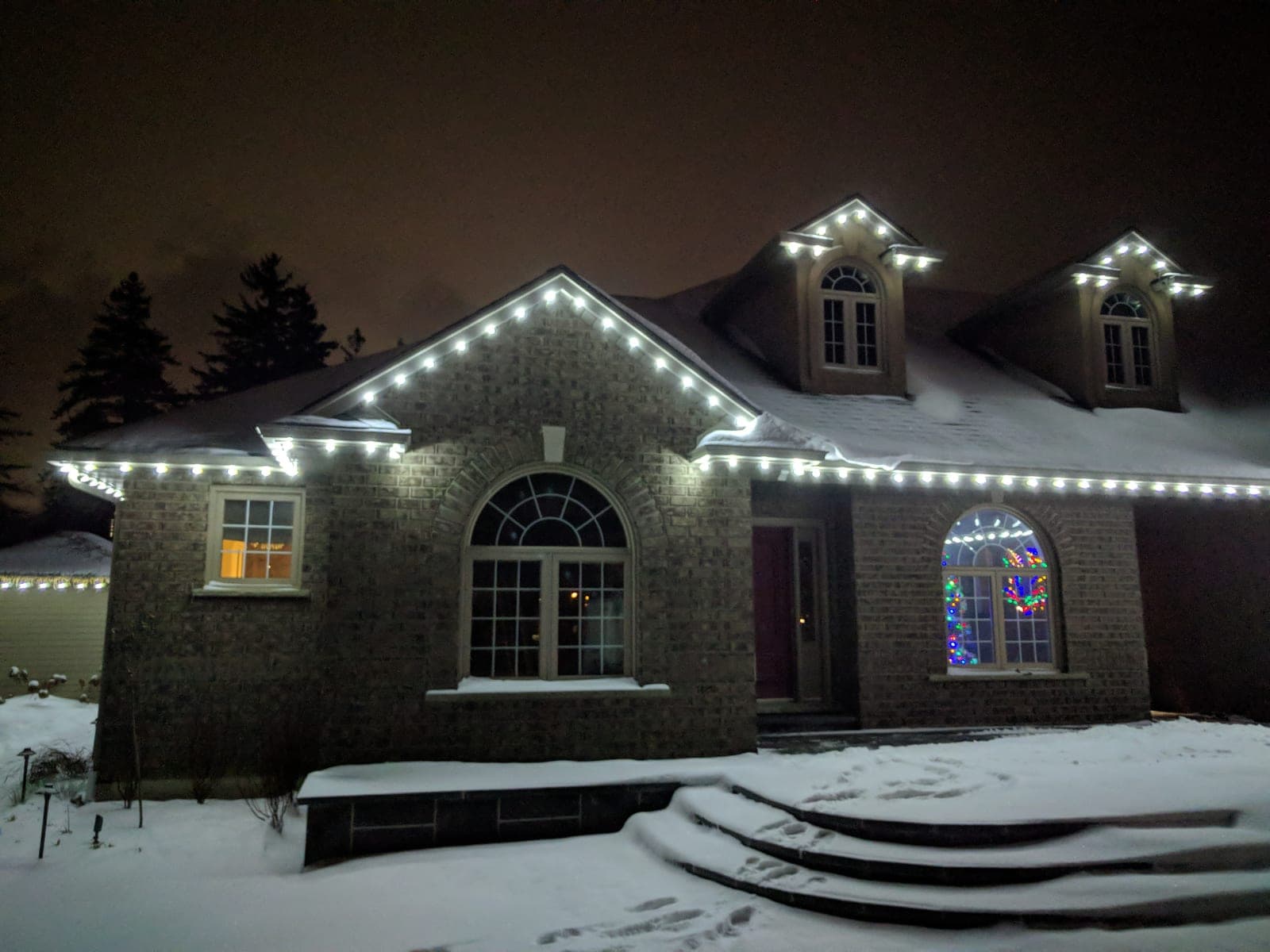 Grey brick home with white roofline lights and colourful lit wreaths glowing in the arched windows